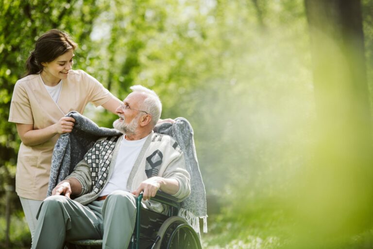 Caregiver helping an older adult in a wheelchair stay warm outdoors by placing a blanket around their shoulders.